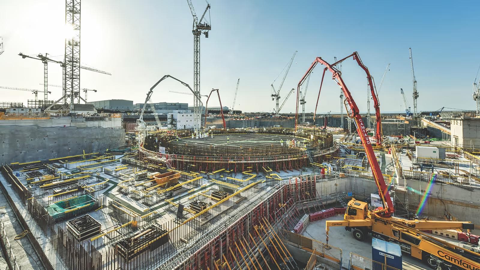 A large-scale construction site under a clear sky, showing the circular foundation of a major structure surrounded by dense rebar, formwork, and scaffolding, with numerous tower cranes and red concrete pump trucks (branded "Camfaud") working across the site.