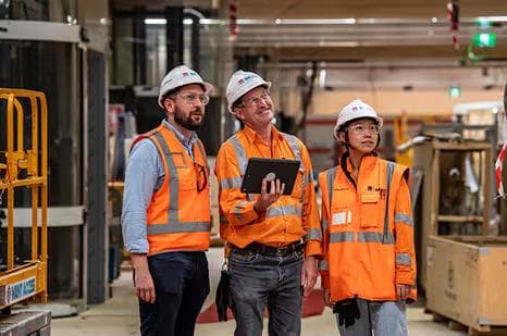 Three construction workers in orange high-visibility gear and white hard hats standing inside an industrial facility, one holding a tablet, looking up at something off-camera.
